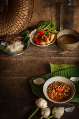 Thai Northern Style Pork and Tomato Chili Relish, nam prik ong in white bowl on wood table there are side dishes of fresh vegetables, cooked rice, water bowl and flower placed around.