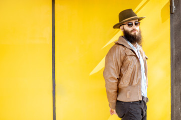 Portrait of a stylish bearded man dressed in jacket and hat on the bright yellow background outdoors