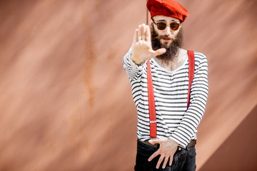 Portrait of a stylish bearded man dressed in striped shirt, suspenders and hat on the red...