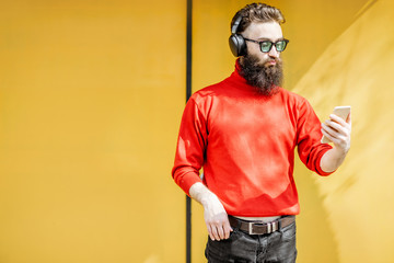 Fototapeta na wymiar Portrait of a stylish man dressed in red sweater enjoying music with headphones and smart phone on the bright yellow background
