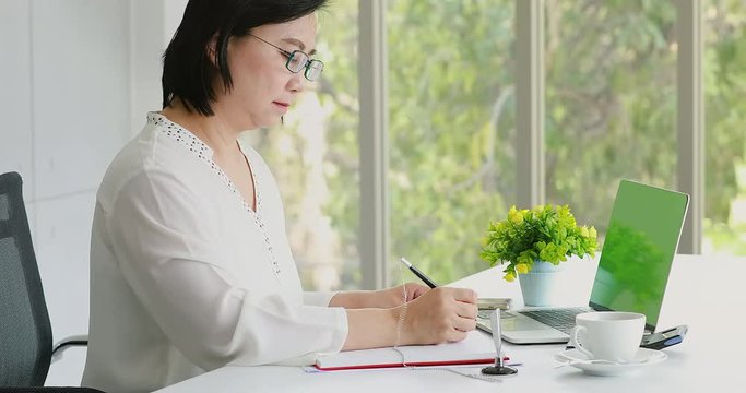 Close up scene video of senior Asian woman writing note on notebook in modern office.