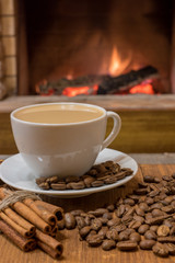 Cup of coffee with milk and coffee beans around the cup, cinnamon sticks before cozy fireplace.