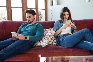 Angry young couple ignoring each other using phone after an argument while sitting on sofa at home.