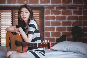 Young beautiful Asian woman is playing acoustic guitar on the bed at her bedroom, vintage warm tone