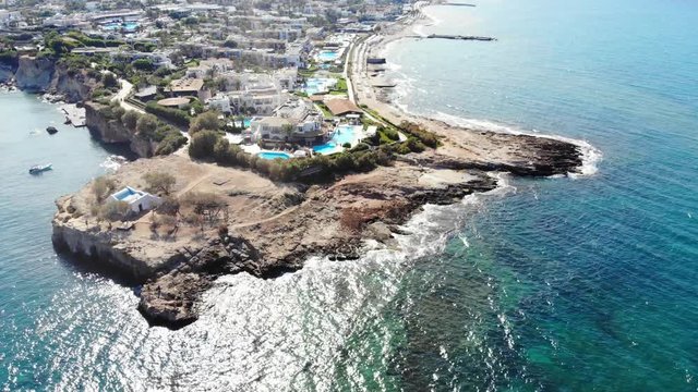 Rocky cape at north coast of Crete Island, hotel buildings and luxury resorts seen on land, aerial shot. Sunny weather, crystal sea water wash rough stony beach