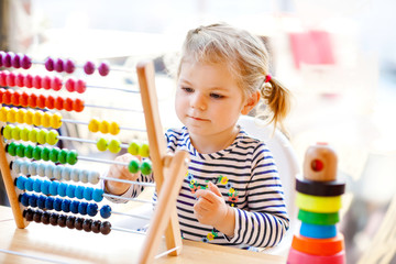 Adorable cute beautiful little toddler girl playing with educational wooden rainbow toy pyramid and counter abacus. Healthy happy baby learning to count and colors, indoors on sunny day.
