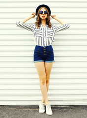 Stylish young woman model in black round hat, shorts, white striped shirt posing on white wall background