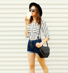 Fototapeta premium Pretty young woman walking with coffee cup in black round hat, shorts, white striped shirt on white wall background
