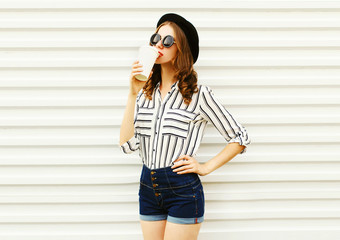 Pretty young woman with coffee cup in black round hat, shorts, white striped shirt on white wall background