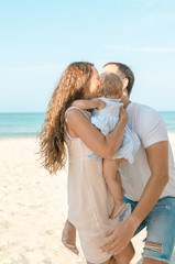 Mother and father holding on hands their infant daughter at the sunny beach background.