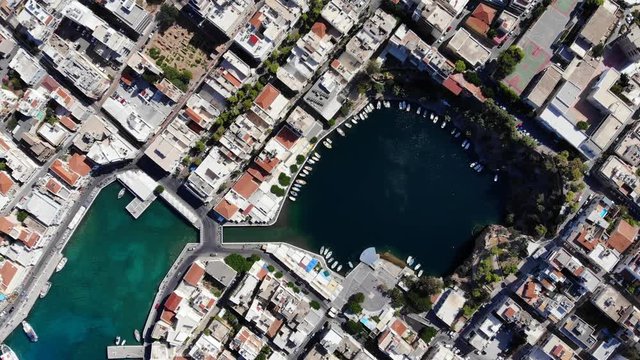 Top aerial view of famous Voulismeni lake at Crete, surround with city buildings. Nice perspective of interesting place, small boats along shore on water, dense build Agios Nikolaos town area around