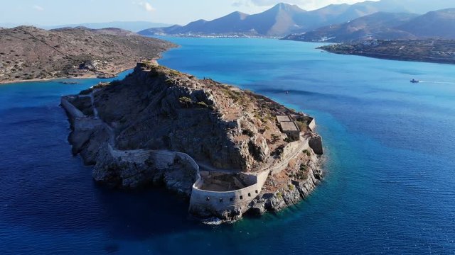 Spinalonga fortress on stony islet, famous landmark of Crete island, aerial shot at sunny evening. Rampart wall on bank of isle, small old buildings ruins on slopes. Clear sea waters around.