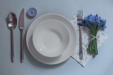 table setting on a blue background, space for one person, two white plates, blue flowers in a bouquet, cutlery, spring table, purple candle