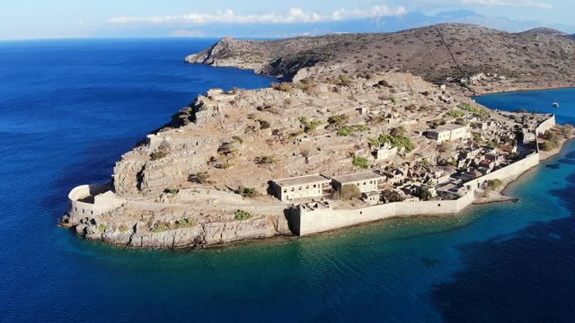 Aerial orbiting shot of Spinalonga isle, interesting historical site at Crete island. Former fortifications, old buildings ruins seen on hill, rampart wall on shore. Sea water surround small island