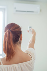 Fototapeta premium Red-haired woman's hand turning on the air conditioner. Red-haired woman stands back and holds a remote control air conditioner at home. Air conditioning in the apartment