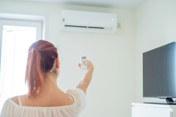 Red-haired woman's hand turning on the air conditioner. Red-haired woman stands back and holds a remote control air conditioner at home. Air conditioning in the apartment