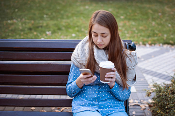 Young and attractive girl sitting on a bench in autumn park and drinking coffee