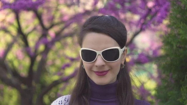 Attractive European girl young woman with long brown hair brunette in white purple clothes in Park wearing white sunglasses smiling, close up portrait, slow motion