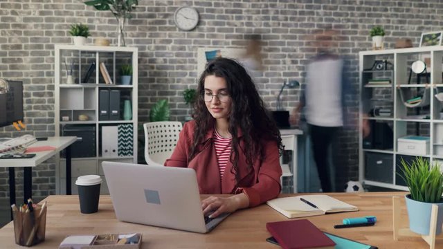 Zoom out time-lapse of beautiful young lady using modern laptop in shared office room focused on job while men and women are moving around. People and work concept.