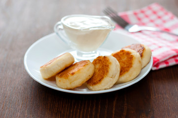 Cheese pancakes with sour cream on a white plate on a pink napkin with a fork, side view. Can be used for menu design.