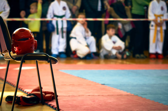 Red Taekwondo helmet on a chair near the tatami for competition. Athletes in the background.