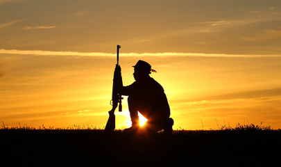 Fototapeta premium Silhouette of a man with a rifle on the background of a beautiful sunset, the boy shoots a gun