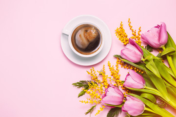 Obraz na płótnie Canvas Pink tulips and mimosa flowers with cup of coffee on pink trendy background. Spring greetings. Flat lay, top view