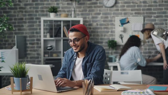 Cheerful young man hipster wearing hat and glasses is using laptop typing working in modern loft style office enjoying modern technology. Youth and work concept.