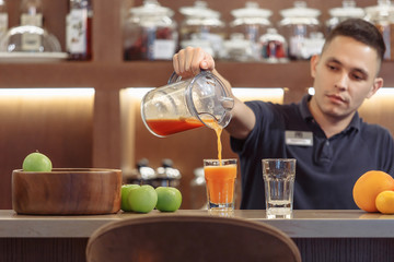 Young handsome barman pours fruit juice standing behind the bar in the evening cafe bar. The concept of non-alcoholic fresh juices
