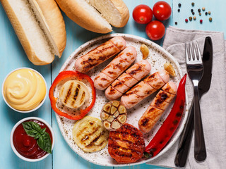 Top view of chicken homemade sausages, buns bread, sauces ketchup and mustard on blue wooden background. Grilled sausages and grilled vegetables in craft trendy plate. Homemade hotdogs.