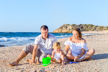 Happy family of three - pregnant mother, father and daughter having fun, playing with sand and shells on the beach. Family vacation, travel concept. Selective focus. Copy space.