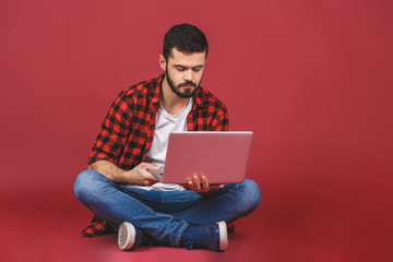 Portrait of a happy young man in casual holding laptop computer while sitting on a floor isolated over red background.
