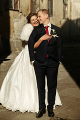 happy gorgeous bride and stylish handsome groom  hugging and laughing on background of old building in sunlight