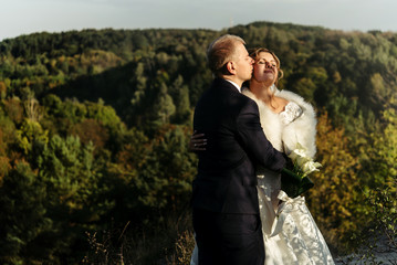 happy gorgeous bride and stylish groom hugging with gentle and kissing on background of sunny trees forest, space for text