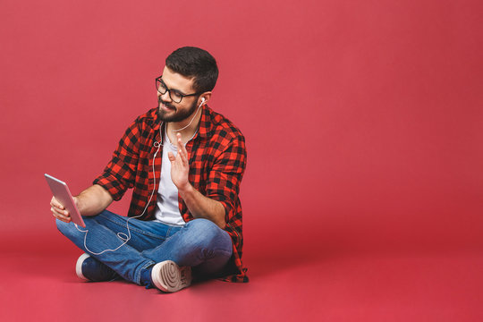Portrait of young man sitting on the floor listening to music with tablet computer isolated over red background.