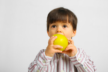 Small caucasian boy with dark hair, big dark eyes, wearing striped shirt is eating, biting big green apple, isolated on white background