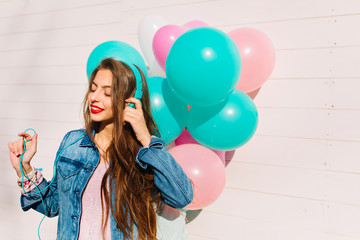 Obraz na płótnie Canvas Close-up portrait of long-haired brunette girl in stylish denim jacket going to birthday party. Attractive young woman posing with bright balloons while listening music in headphones with closed eyes