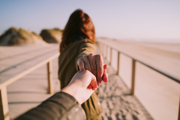 A couple is holding hands in the beach. One of the persons is following the other while walking. Selective focus. Close up.