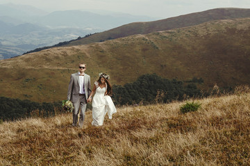 happy gorgeous bride and groom walking  in sun light holding hands, boho wedding couple, luxury ceremony at mountains with amazing view, space for text