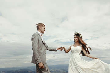 Fototapeta na wymiar gorgeous bride and groom holding hands and dancing at sky and clouds, moment of true happiness, luxury ceremony at mountains with amazing view, space for text