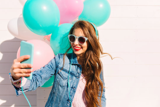 Charming birthday girl in sunglasses accepts congratulations on video communication using a smartphone. Gorgeous young woman in headphones and denim jacket making selfie with balloons at the party