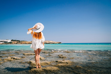 Red-haired female in white dress stands with his back to camera on shores of Mediterranean Sea