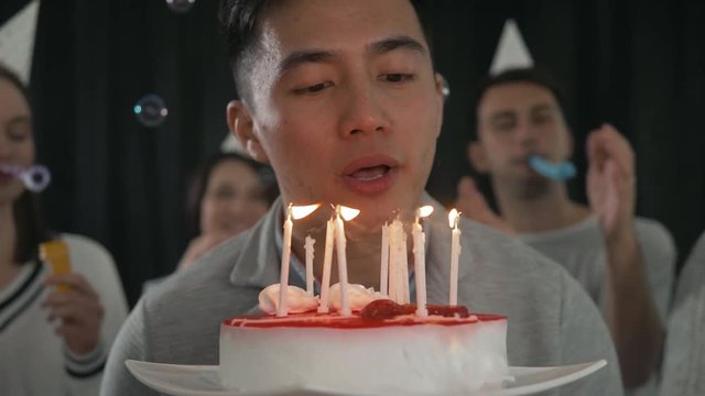 A group of young people celebrate the birthday of one of their friends. A young asian man is blowing candles on the cake. Sparkles in hands happy friends