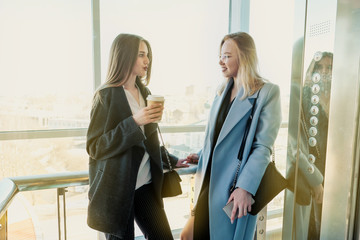 Two beautiful girls, a blonde and a brunette, are wearing a coat in a glass elevator with a window. Shopping center or office in the city. © spaskov