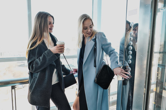 Two beautiful girls, a blonde and a brunette, are wearing a coat in a glass elevator with a window. Shopping center or office in the city.