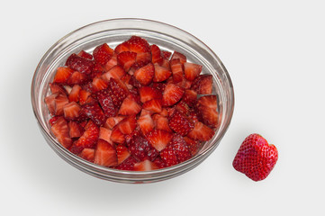 A glass bowl  with chopped strawberries and one outside, isolated on white background seen from up close