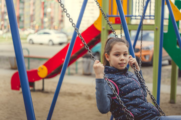 Girl swinging on colourful playground Teenage girl in warm clothes having fun on swing and looking at camera