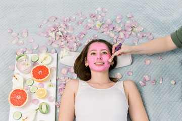 Fototapeta na wymiar Spa therapy for young smiling woman receiving facial mask at beauty salon - indoors. Pretty girl with pink facial mask on face lying on bed, fruits and flowers around her