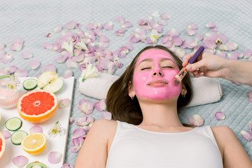 Obraz na płótnie Canvas Woman with pink clay facial mask in beauty spa. In background flower petals and fresh fruits, grapefruit slices, apple, lemon