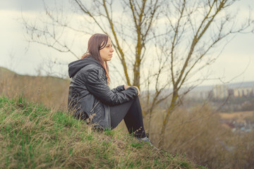 Young woman sitting on green hill Side view of young dreamy female sitting on altitude and looking at suburb in spring day